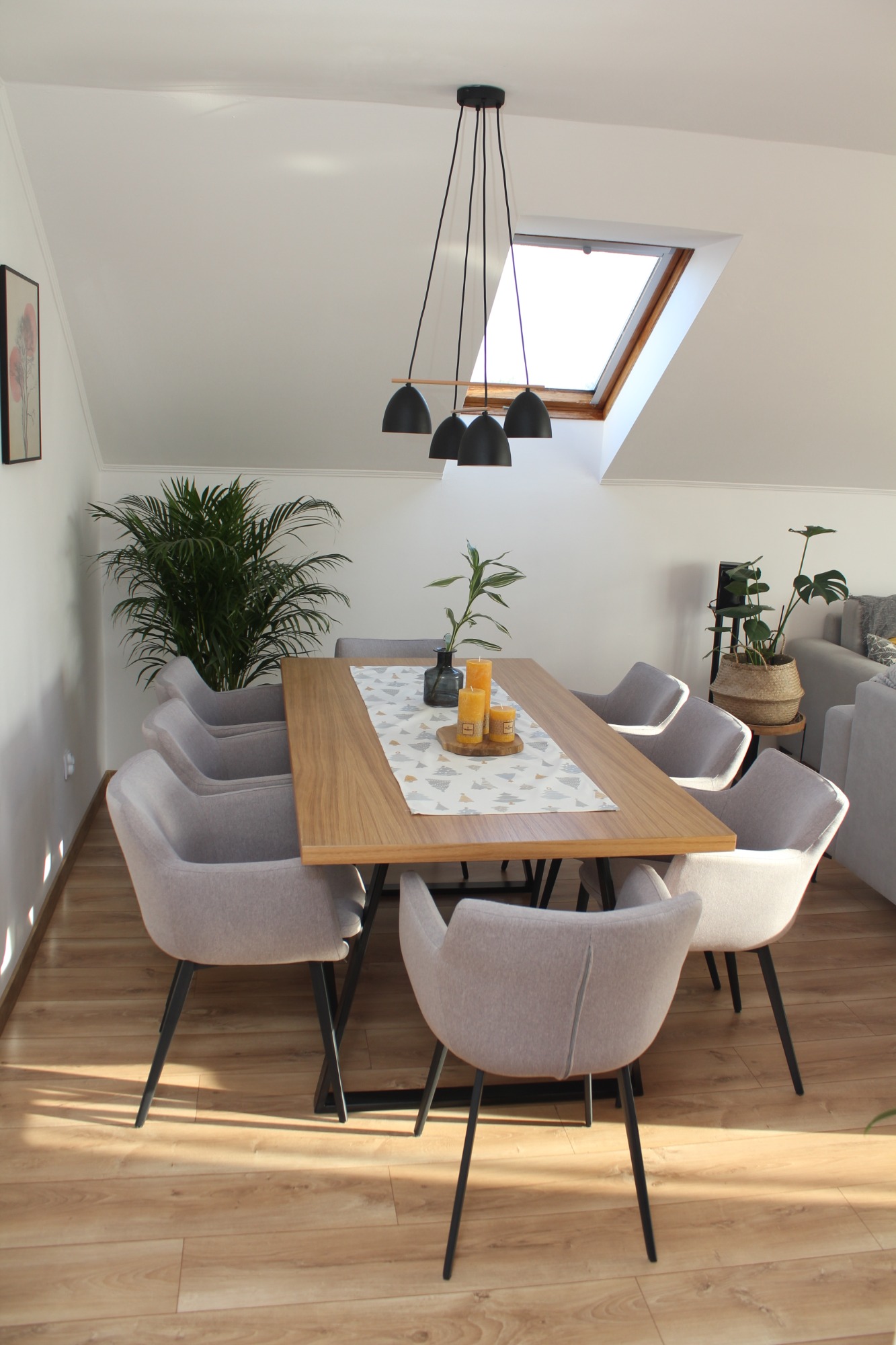Jasna jadalnia na poddaszu z drewnianym stołem, szarymi krzesłami na czarnych nogach, oświetlona przez okno dachowe i wiszące lampy, z roślinami doniczkowymi jako elementami dekoracyjnymi.