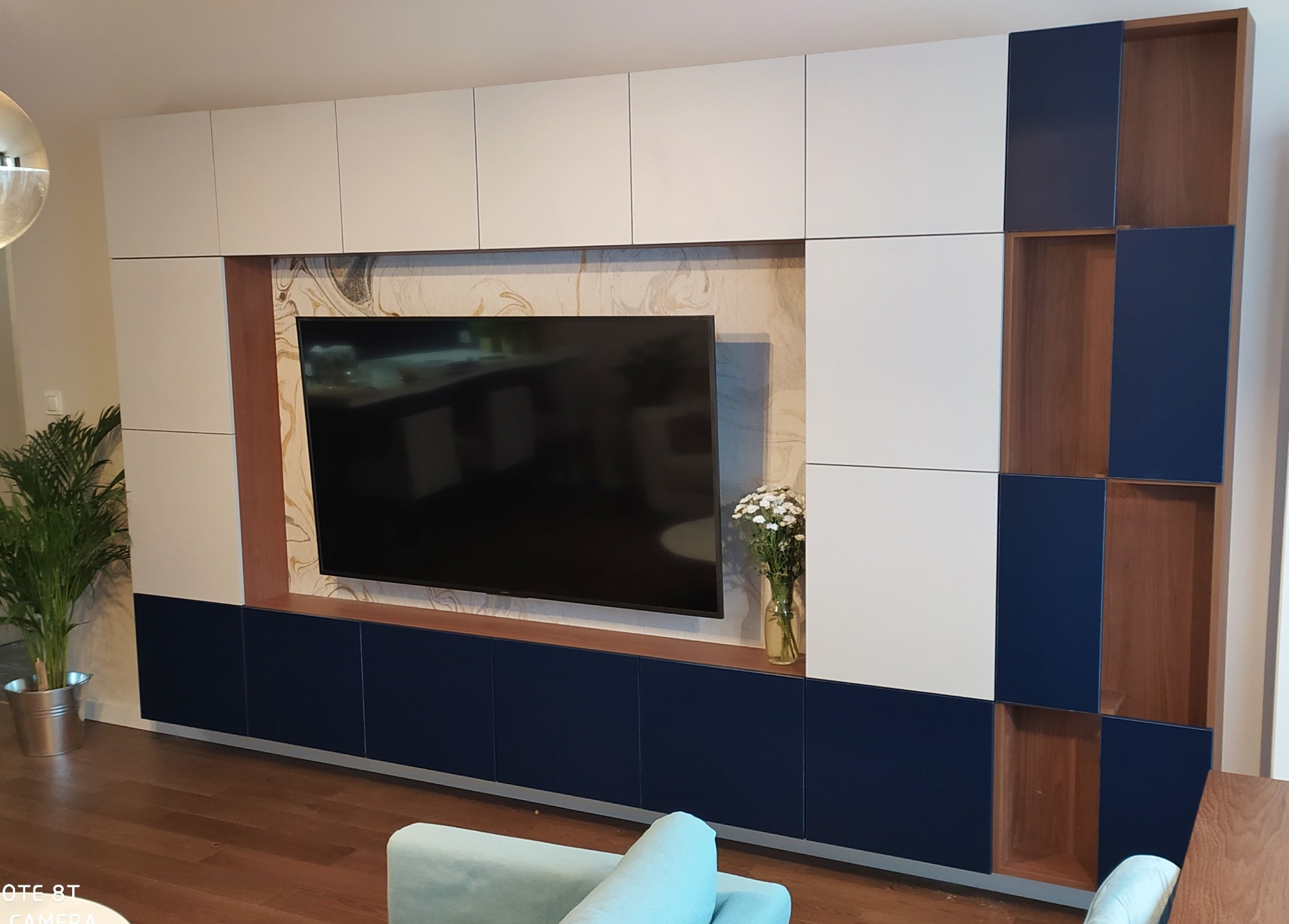 Nowoczesna zabudowa ściany w salonie: białe i granatowe szafki z drewnianymi akcentami, telewizor na tle marmurowej ściany, kwiaty w wazonie.