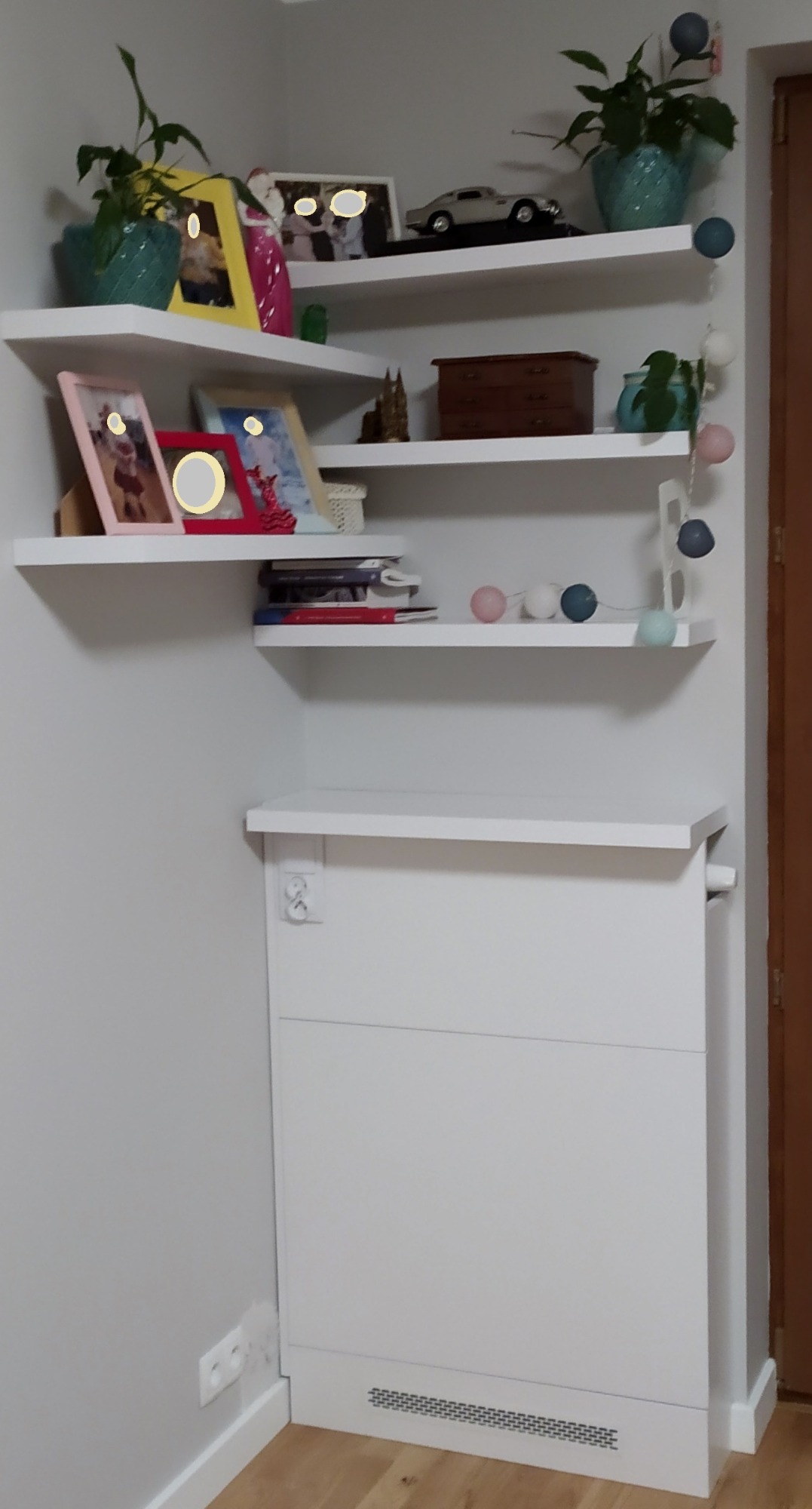 Białe półki narożne z dekoracjami: ramki ze zdjęciami, rośliny w doniczkach, drewniana szkatułka, model samochodu, girlanda z kulkami, nad zabudowanym grzejnikiem.