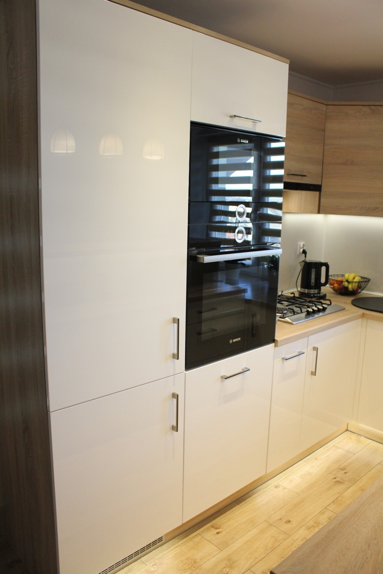 Biała, nowoczesna zabudowa kuchenna z piekarnikiem Bosch, jasny drewniany blat i podłoga, widoczne odbicia lamp w lakierowanych frontach.