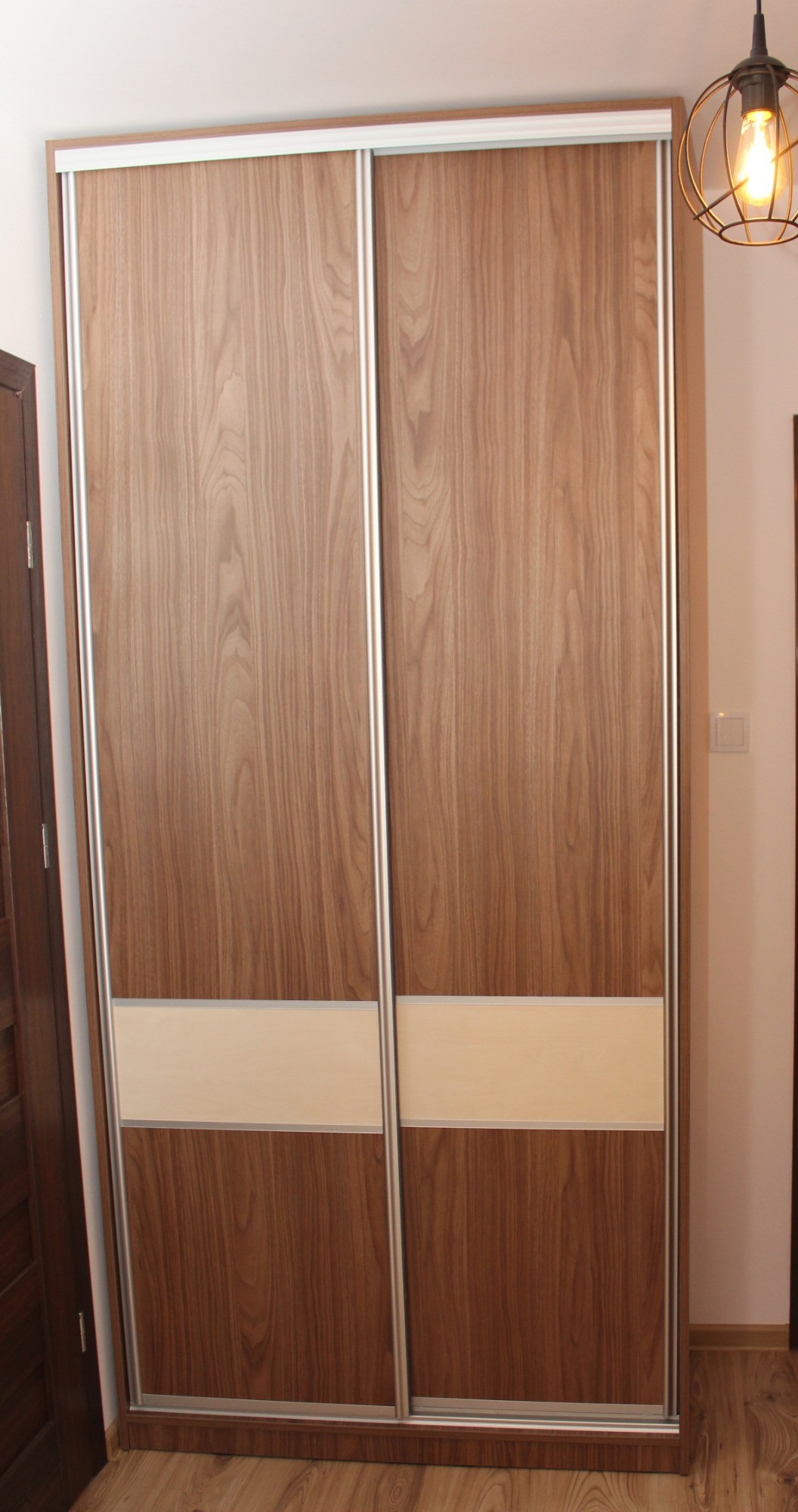 Dwudrzwiowa szafa przesuwna z okleiną drewnopodobną i jasnym pasem na dole, oświetlona żarówką Edisona w metalowej klatce.