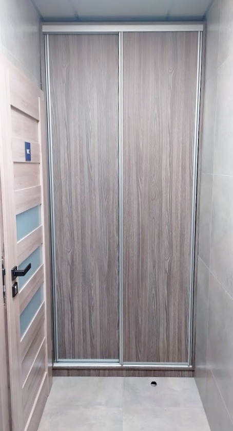 Zamknięta szafa przesuwna z drewnopodobnymi frontami w odcieniach szarości i beżu, z widocznymi aluminiowymi profilami, umieszczona we wnęce obok otwartych drzwi wewnętrznych z przeszkleniami.