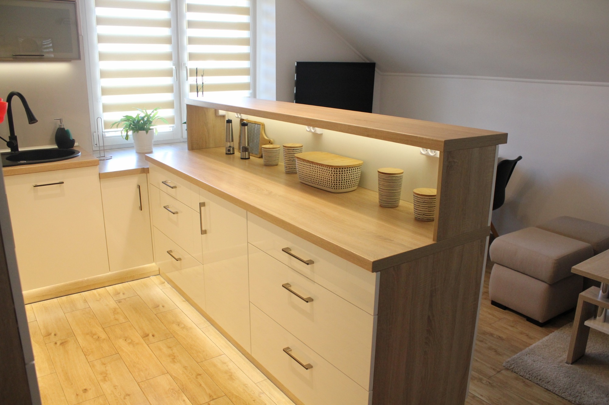 Białe szafki kuchenne z drewnianym blatem i podświetleniem LED, widok na okno z żaluzjami poziomymi i dekoracyjne akcesoria na blacie.