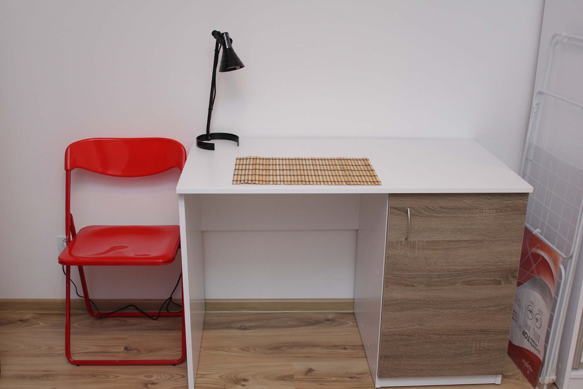 Białe biurko z drewnianym frontem szafki, czarna lampka biurkowa, czerwone składane krzesło, bambusowa podkładka na blacie, suszarka na pranie w tle.