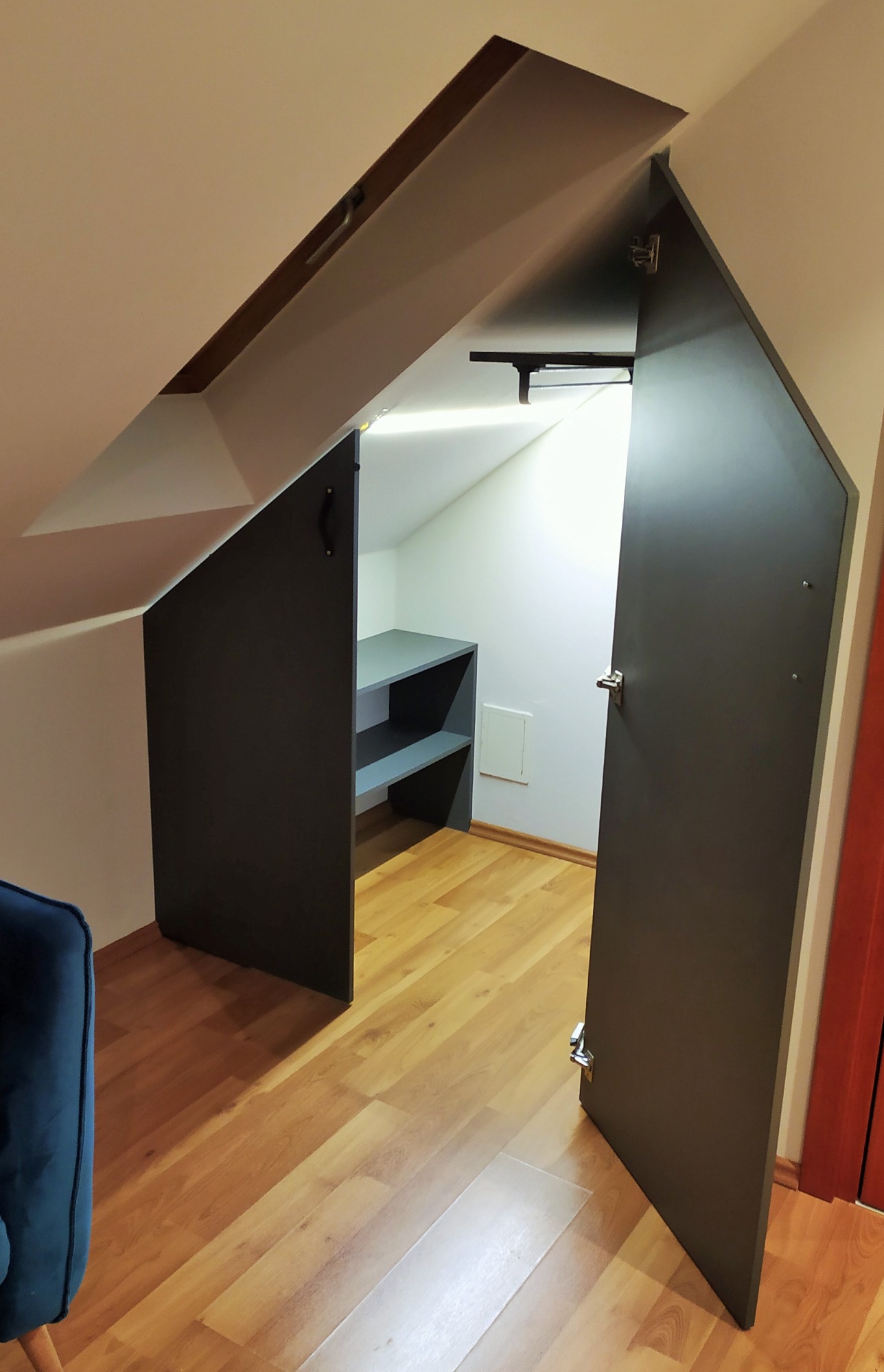 Otwarta, ciemna szafa wnękowa z szarymi półkami wewnątrz, zamontowana na poddaszu, widoczna na tle drewnianej podłogi.