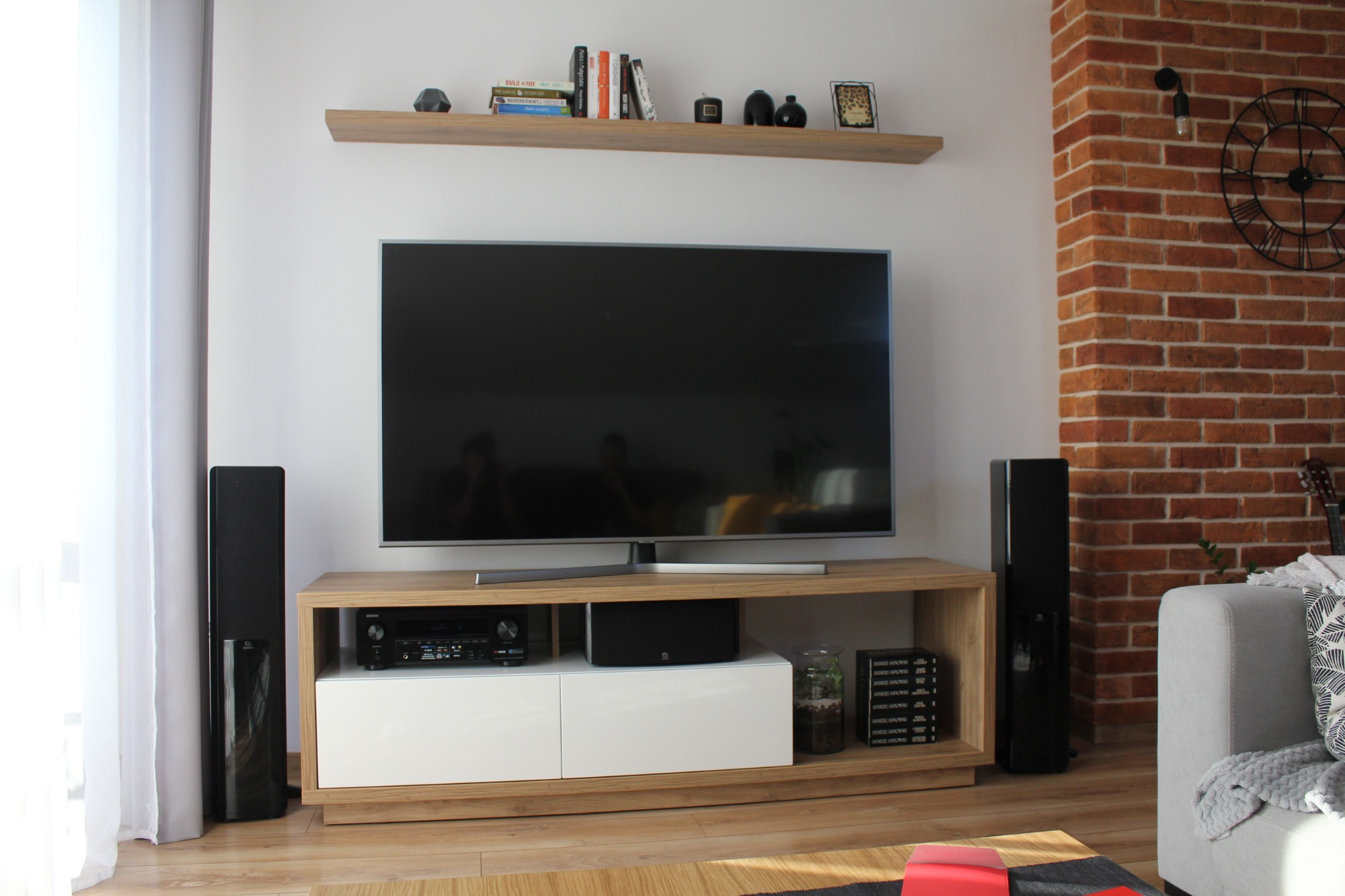Nowoczesny salon z telewizorem na drewnianej szafce RTV z białymi frontami, czarne głośniki po bokach, ceglana ściana z dekoracyjnym zegarem w tle.
