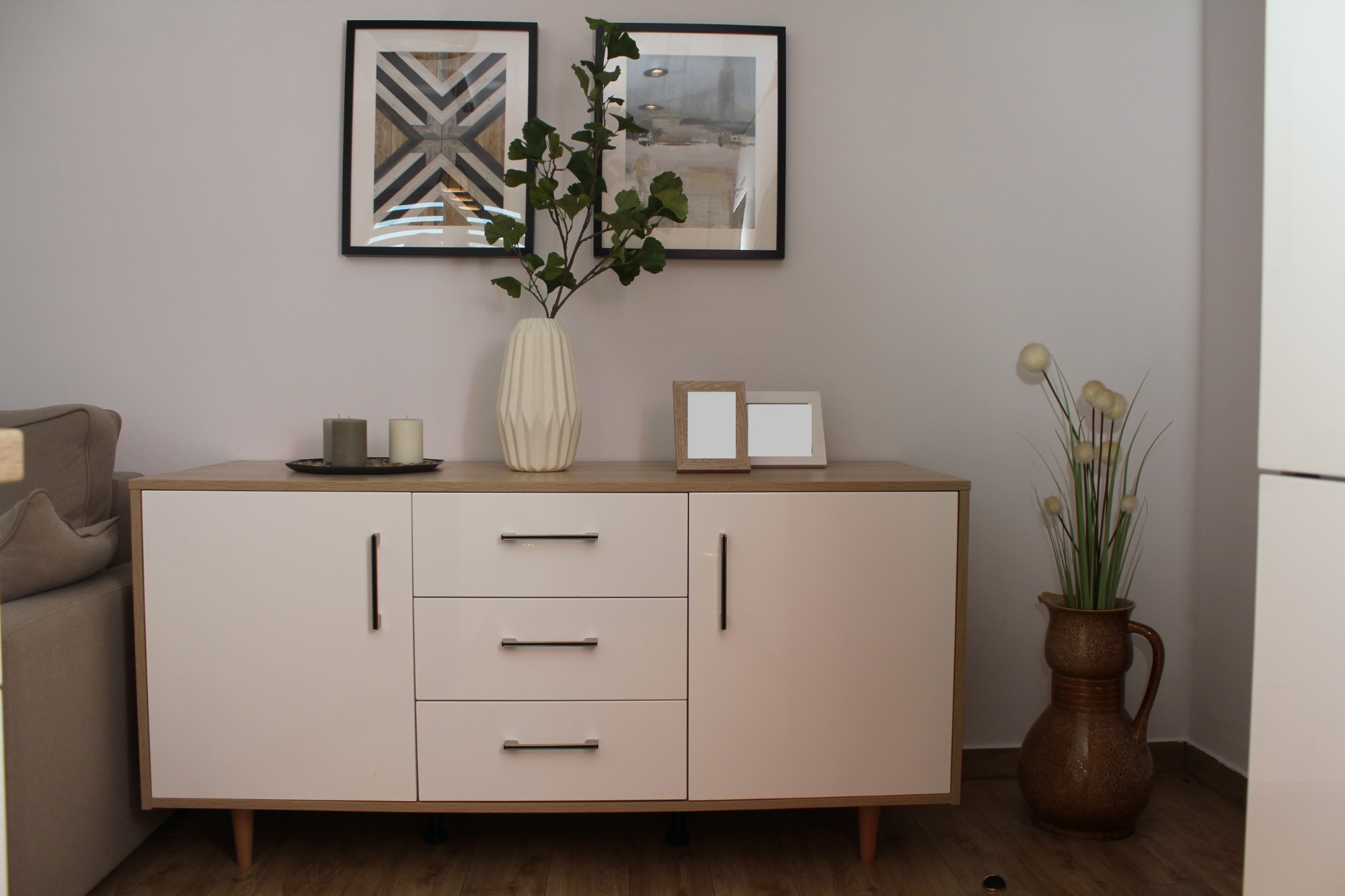 Biała komoda z drewnianym blatem na nóżkach, dekorowana wazonami z roślinami, świecami i ramkami na zdjęcia, na tle szarej ściany z obrazami.