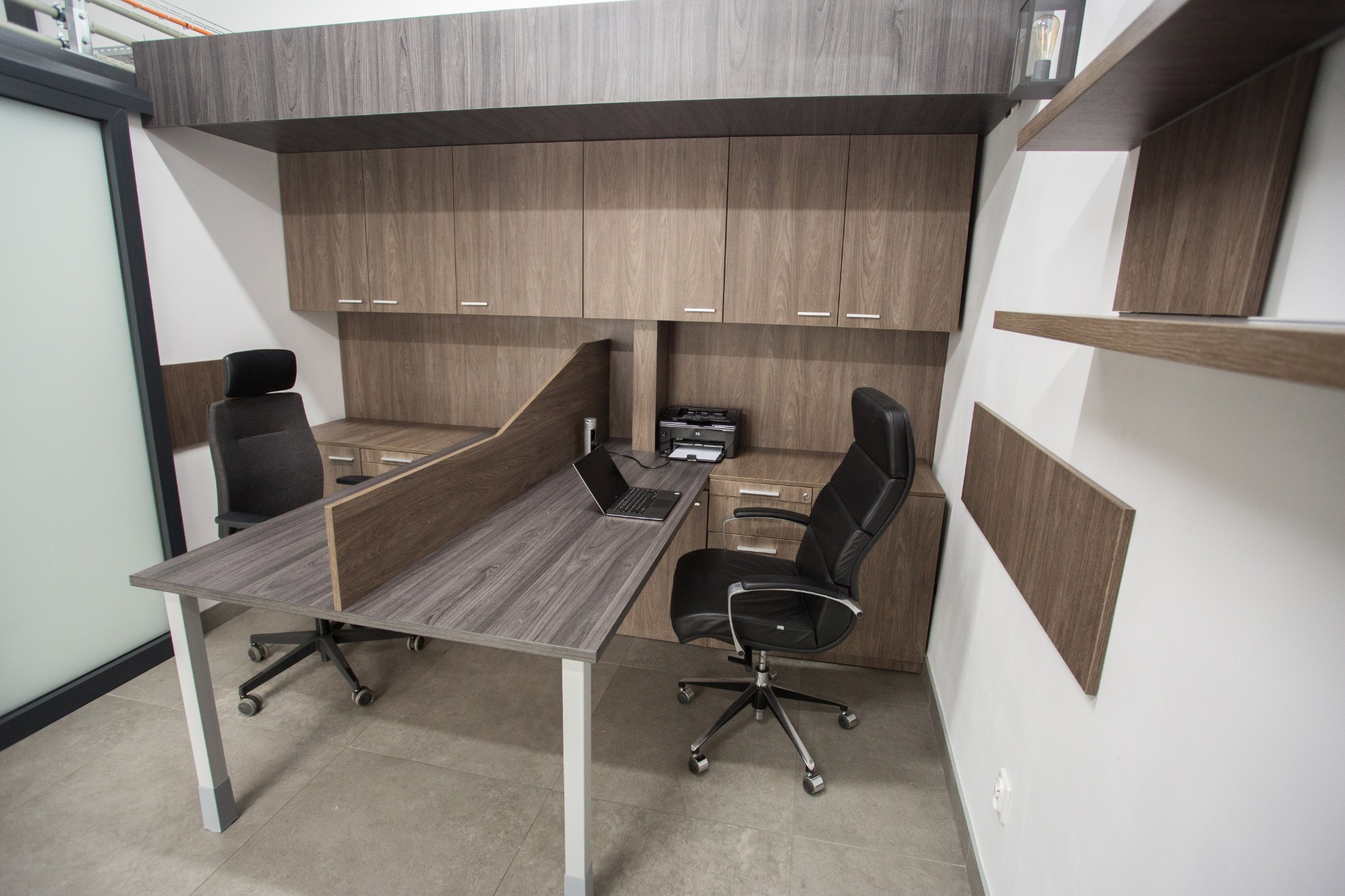 Biuro z dwoma stanowiskami pracy: szare biurka z przegrodami, krzesła biurowe, szafki wiszące z drewnopodobnymi frontami i otwarte półki na ścianie.