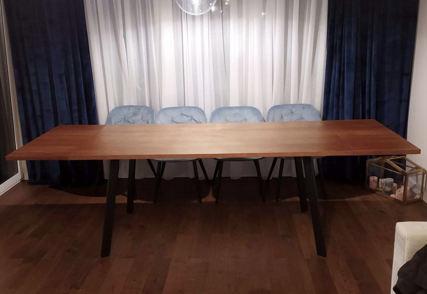 Stół z blatem drewnianym na czarnych nogach, otoczony czterema błękitnymi krzesłami, na tle białych i granatowych zasłon, w mieszkaniu w Krakowie.