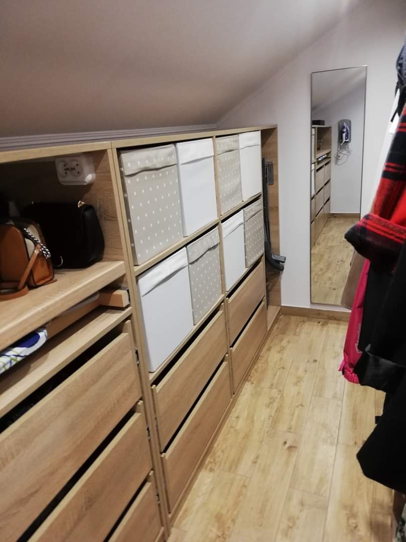 Garderoba na poddaszu ze skośnym sufitem, jasne drewniane szafy z szufladami i pudełkami, lustro odbijające wnętrze szafy, widoczna podłoga z paneli.