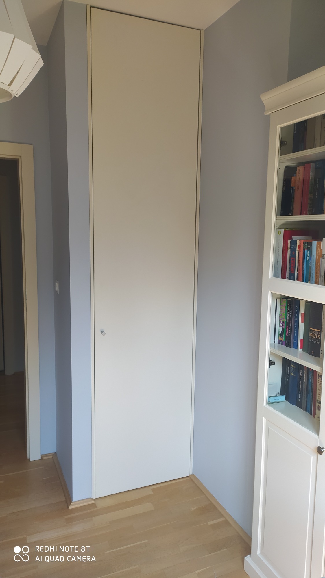 Biała, wbudowana szafa wnękowa z gładkimi frontami, zamontowana od podłogi do sufitu w pokoju z jasnymi ścianami i drewnianą podłogą, widoczna obok białego regału na książki.