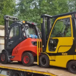 Dwa wózki widłowe, czerwony Linde i żółty Yale, przewożone na platformie ciężarówki z widocznym numerem telefonu, na tle zieleni.