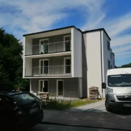 Nowoczesny, dwupiętrowy budynek z szarą elewacją i białymi elementami, z balkonami z balustradami ze stali nierdzewnej i szkła, częściowo zasłonięty przez zaparkowane samochody i stosy materiałów...