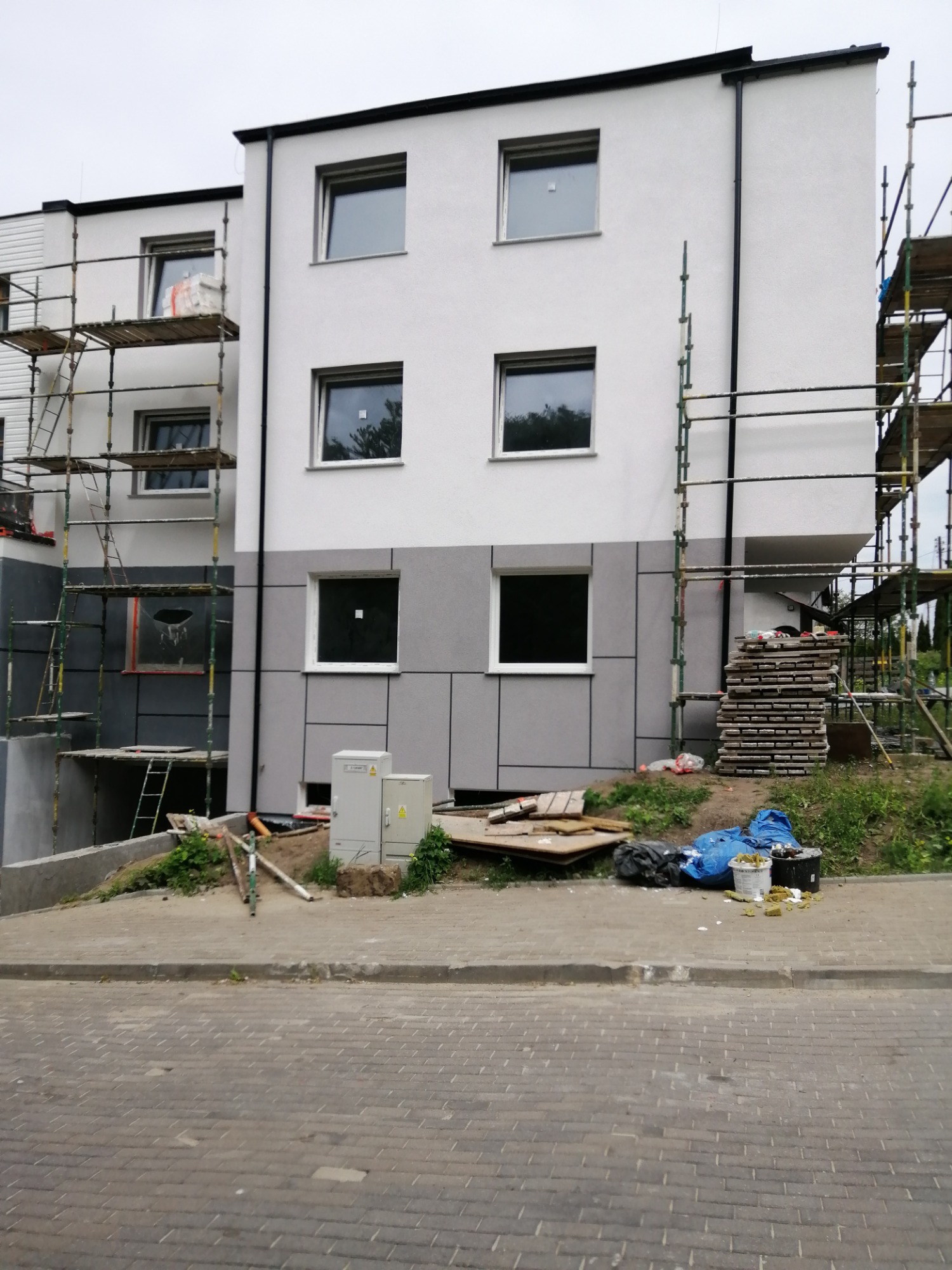 Dwukondygnacyjny budynek w trakcie wykonywania elewacji, widoczne rusztowania, częściowo wykończona elewacja z szarego panelu na dole i białego tynku na górze, elementy ocieplenia leżące na chodniku.