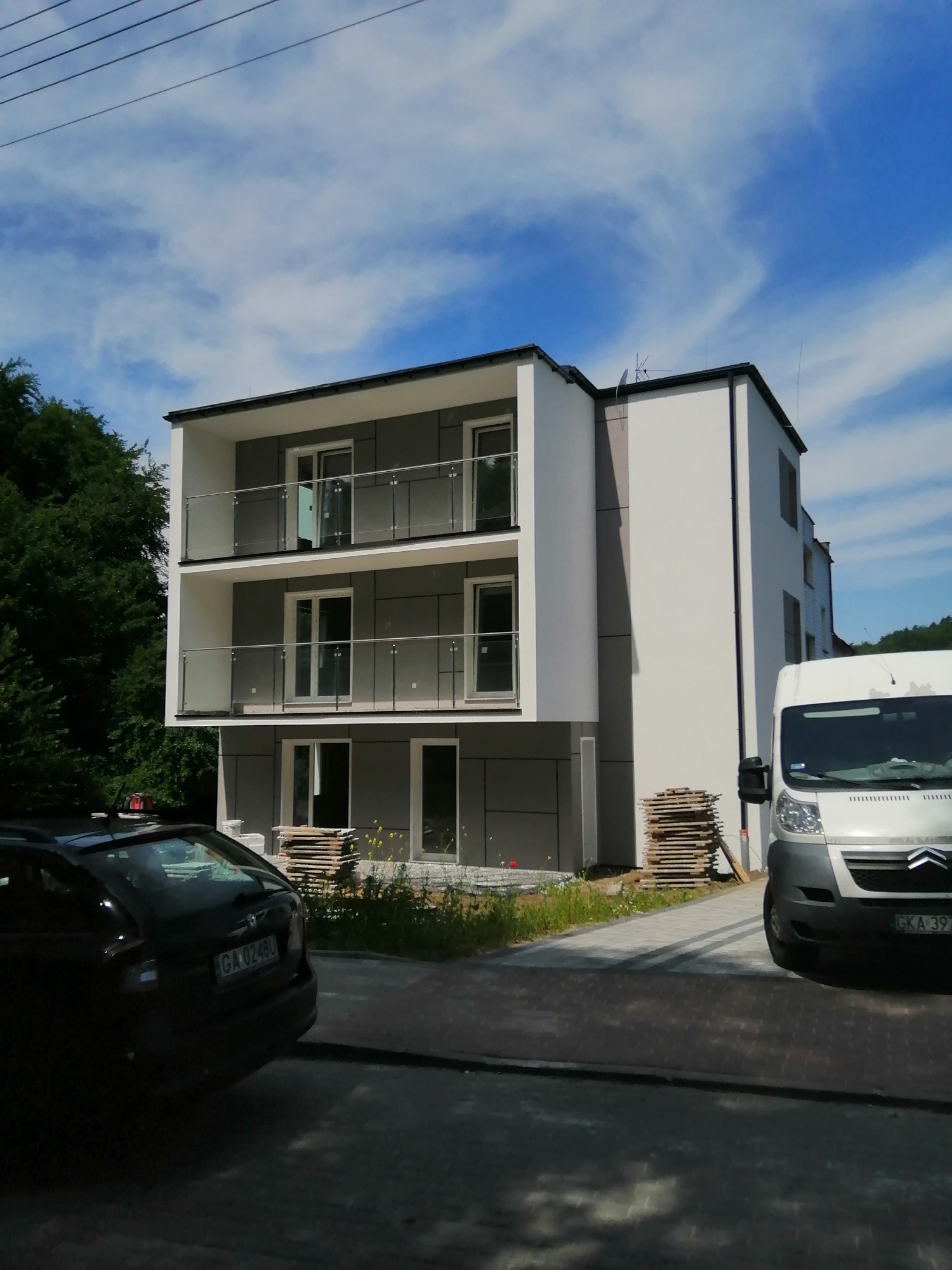 Nowoczesny, dwupiętrowy budynek z szarą elewacją i białymi elementami, z balkonami z balustradami ze stali nierdzewnej i szkła, częściowo zasłonięty przez zaparkowane samochody i stosy materiałów...