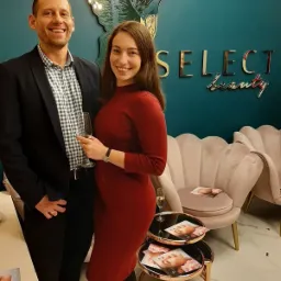 Uśmiechnięta para w eleganckim wnętrzu salonu kosmetycznego, z dekoracją w kształcie kolibra z oświetleniem LED na turkusowej ścianie, logo 'SELECT beauty', filiżanki z szampanem i ulotki...