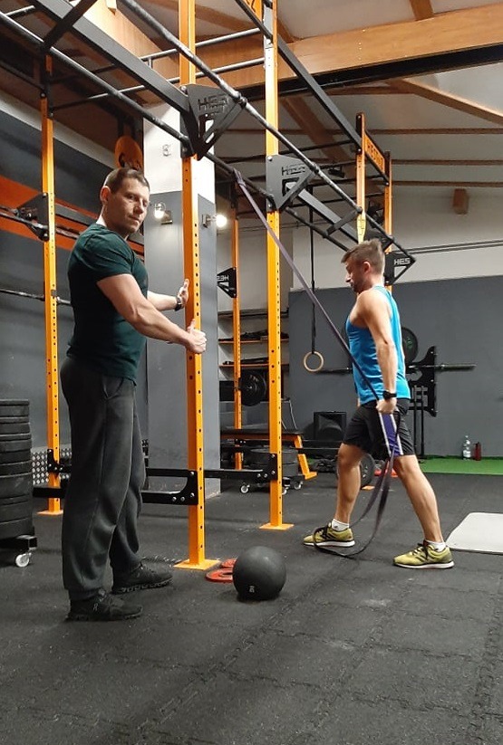 Mężczyzna w niebieskim tank topie ćwiczy z gumą oporową pod nadzorem mężczyzny w zielonej koszulce w siłowni z drabinkami i obciążeniami