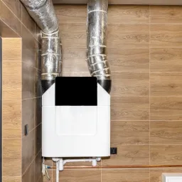 Białe urządzenie z czarnym panelem i srebrnymi rurami wentylacyjnymi na tle brązowych płytek imitujących drewno, obok ciemnych drzwi wejściowych.