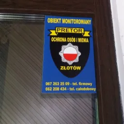 Niebieska naklejka z logo firmy ochroniarskiej Pretor na drzwiach, informująca o monitoringu obiektu w Złotowie, zawierająca numery telefonów kontaktowych.
