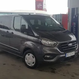 Szary bus Ford Transit Custom z numerem rejestracyjnym NO 8354T, zaparkowany na stacji benzynowej Verwa, z widocznym autobusem w tle.