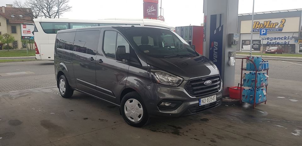 Szary bus Ford Transit Custom z numerem rejestracyjnym NO 8354T, zaparkowany na stacji benzynowej Verwa, z widocznym autobusem w tle.