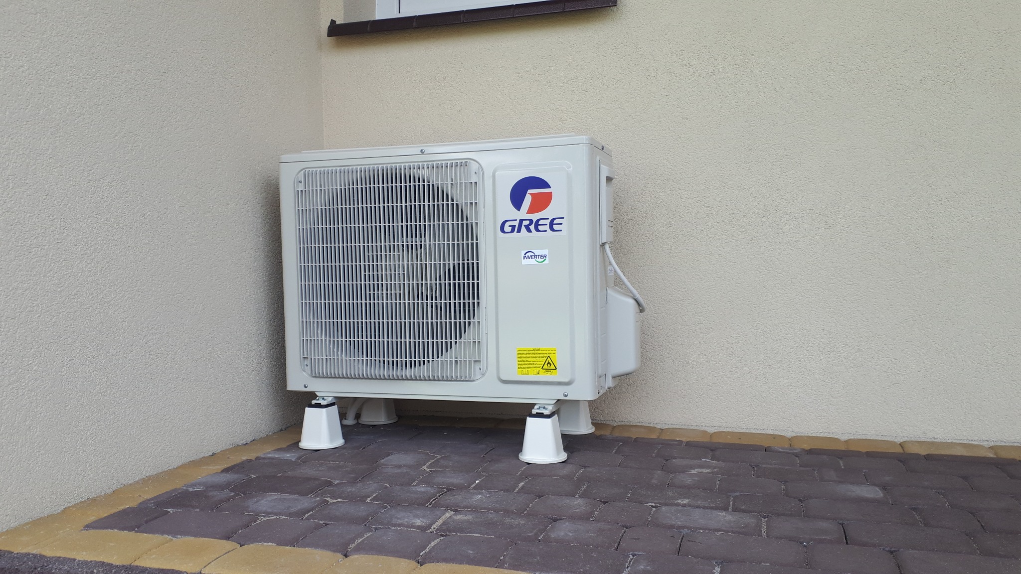 Zewnętrzna jednostka klimatyzacji GREE MUSE 3,5kW zamontowana na białych wspornikach na brukowanym tarasie przy ścianie budynku.
