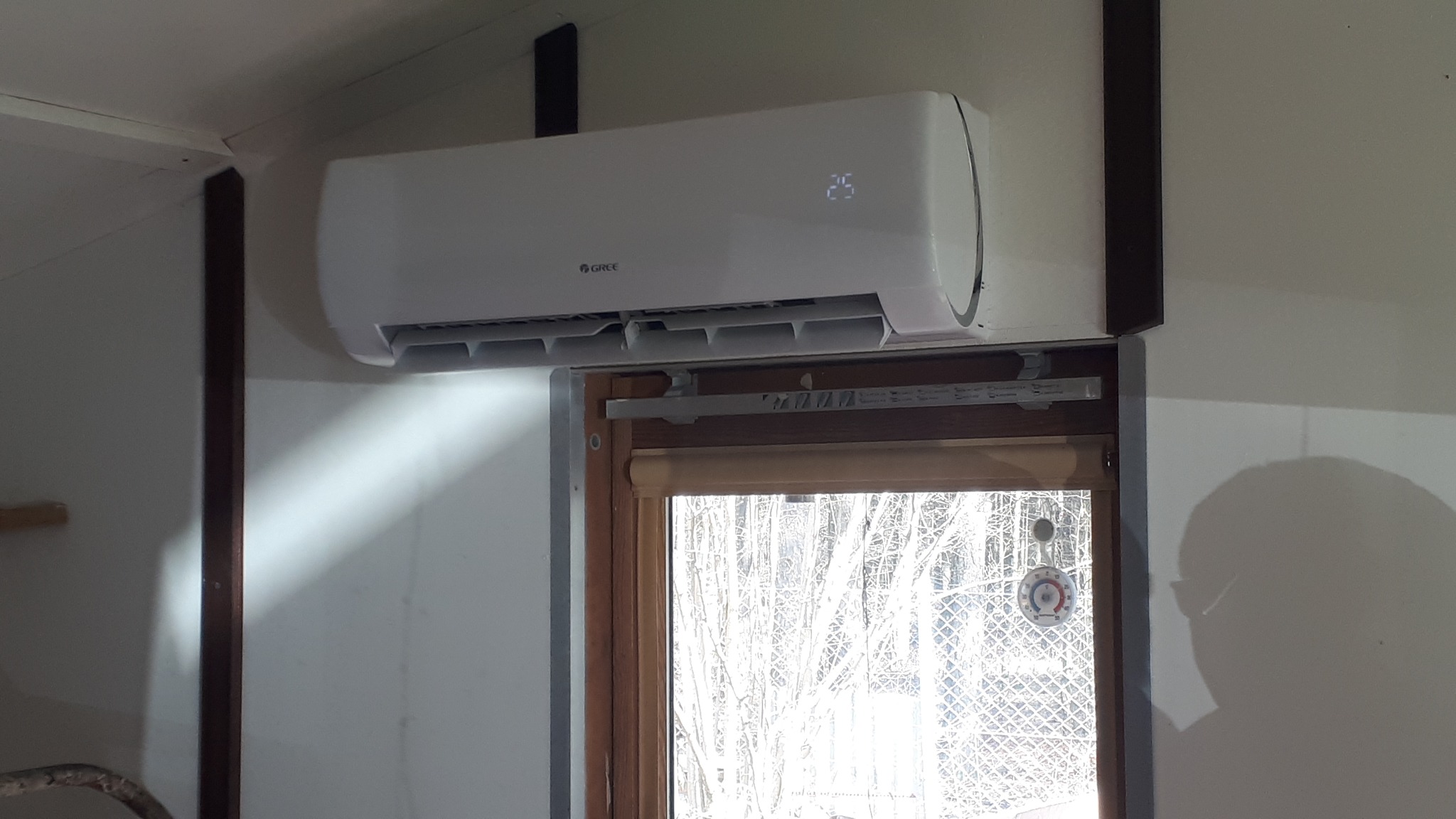 Biała jednostka klimatyzacji GREE zamontowana na ścianie nad oknem z widoczną temperaturą 25 stopni Celsjusza na wyświetlaczu.