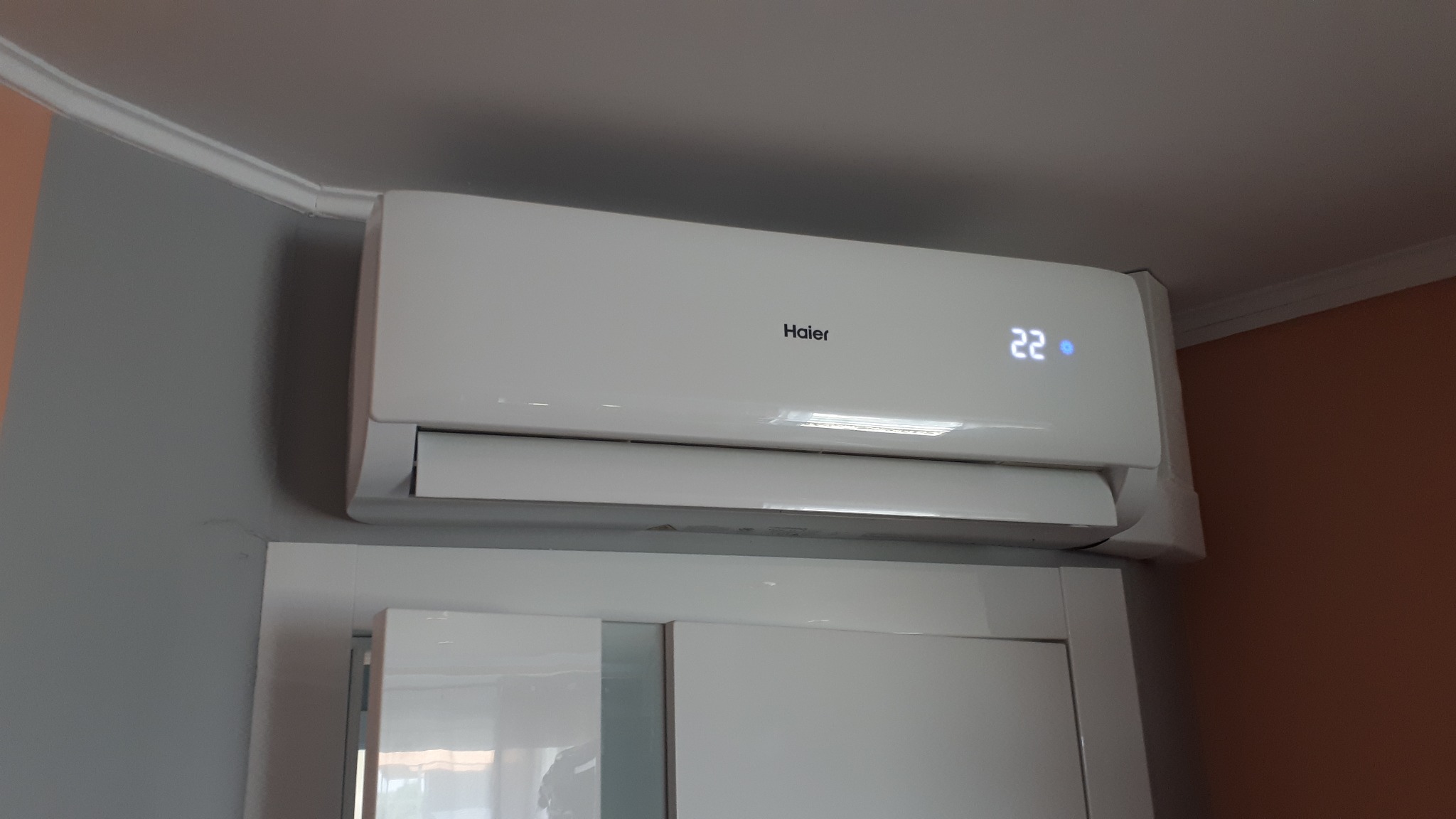 Biała klimatyzacja Haier Tundra z wyświetlaczem temperatury 22 stopnie Celsjusza, zamontowana nad szafką, widoczna na tle szarej i pomarańczowej ściany.