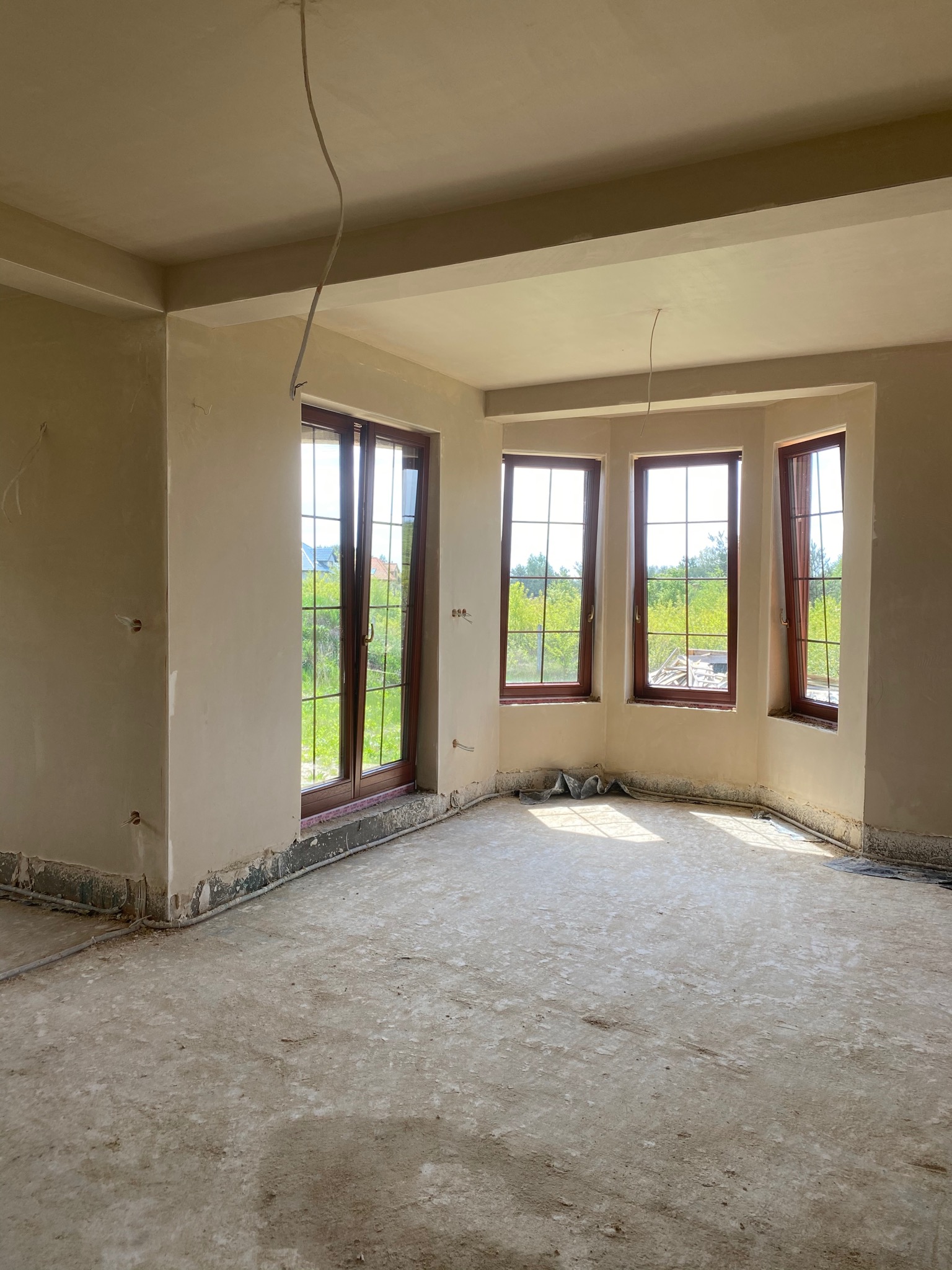 Wnętrze pokoju po nałożeniu tynków, widoczne okna z brązowymi ramami i surowa podłoga, wystające kable ze ściany i sufitu.