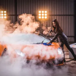 Kobieta w czarnej skórzanej kurtce i spodniach czyści pomarańczowe Lamborghini parą i niebieską ściereczką, obok stoi parownica z napisem Volt, w tle blask lamp.