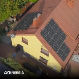 Widok z góry na dom z brązową dachówką i zamontowanymi panelami fotowoltaicznymi na dachu, widoczny również komin i antena satelitarna, na dole logo firmy AZ Energia.
