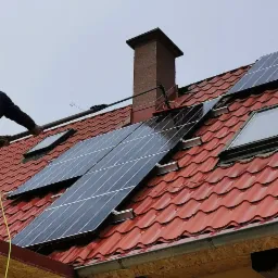 Czyszczenie paneli słonecznych na dachu z czerwonej dachówki, widoczny komin i okno dachowe, osoba używa lancy teleskopowej z dyszą.