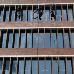 Trzech alpinistów przemysłowych, w kaskach i uprzężach, zawieszonych na linach, czyści okna na wysokościach nowoczesnego biurowca z cegły.