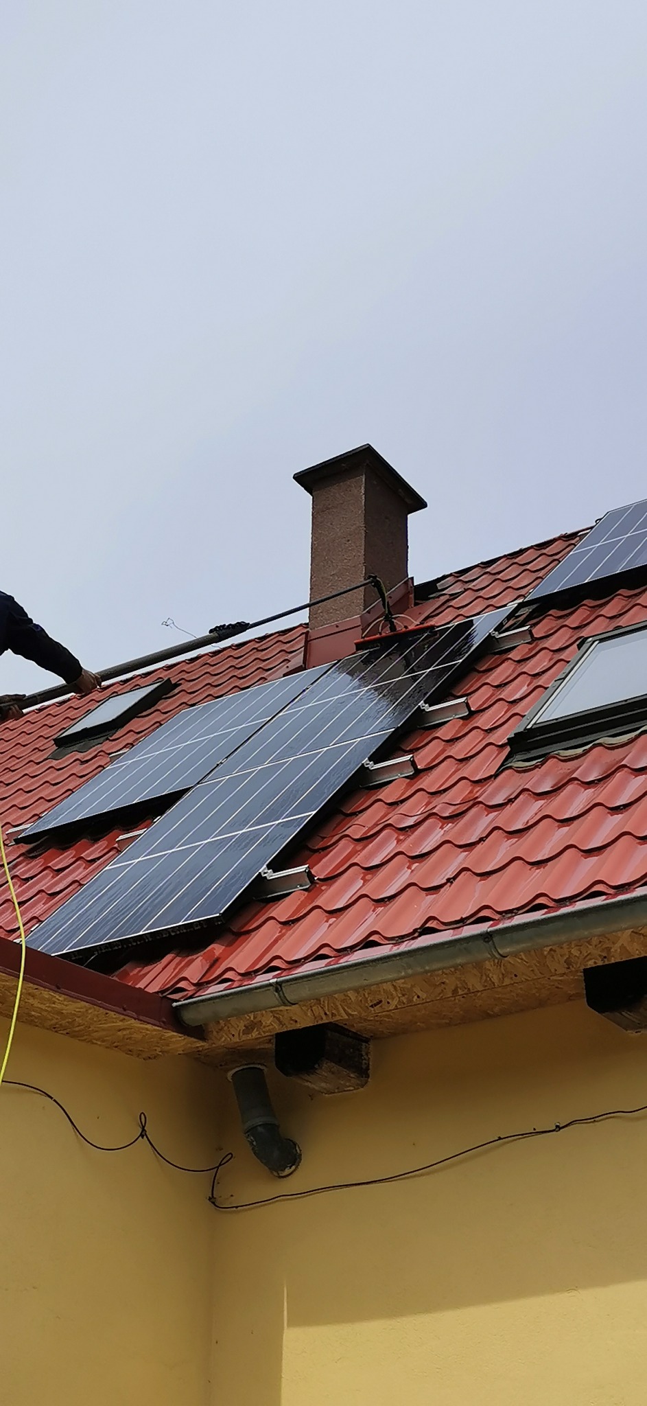 Czyszczenie paneli słonecznych na dachu z czerwonej dachówki, widoczny komin i okno dachowe, osoba używa lancy teleskopowej z dyszą.