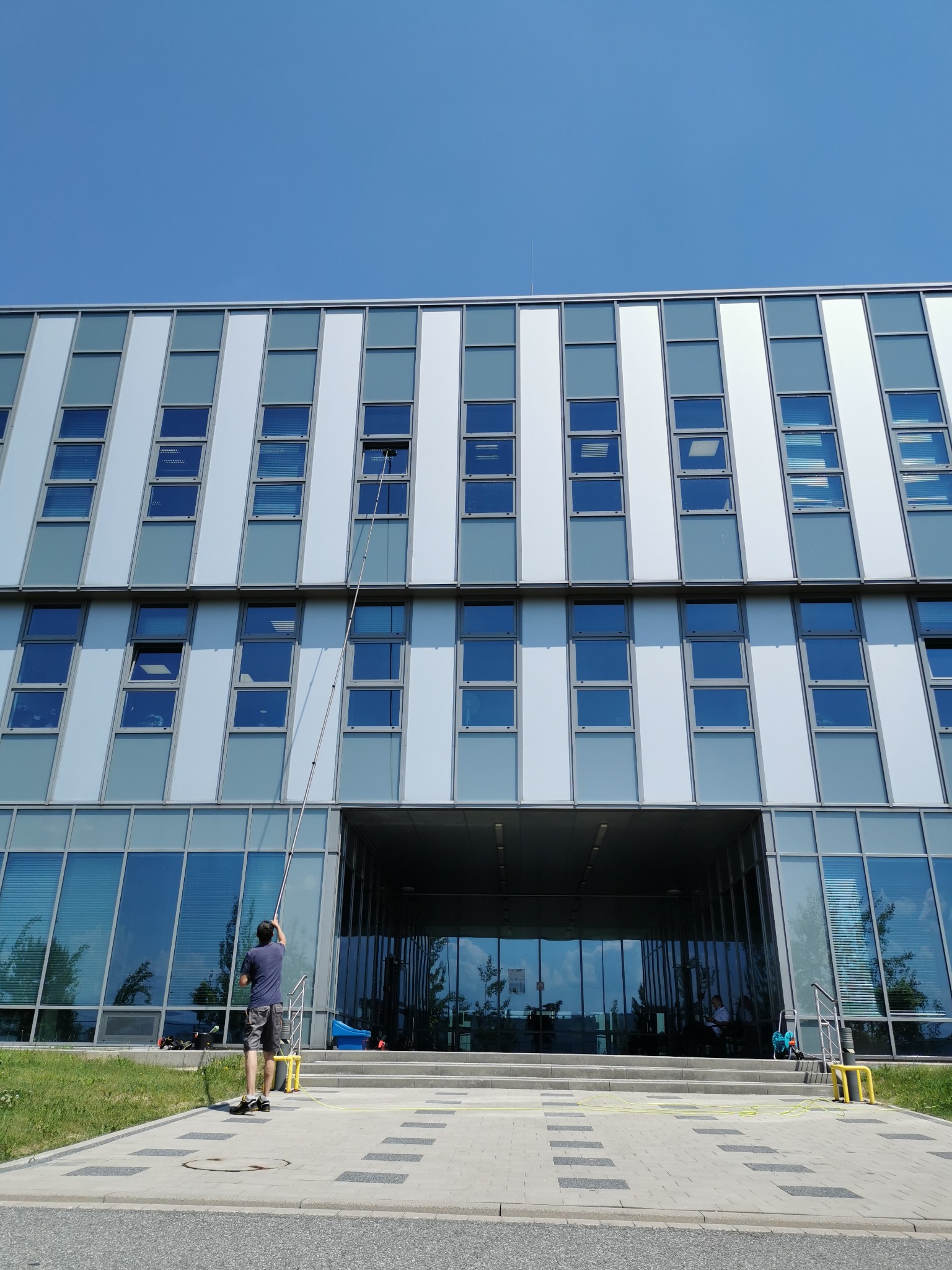 Pracownik z ziemi myje okna na wysokościach przy użyciu teleskopowego kija z szczotką na tle nowoczesnego biurowca z dużymi oknami.