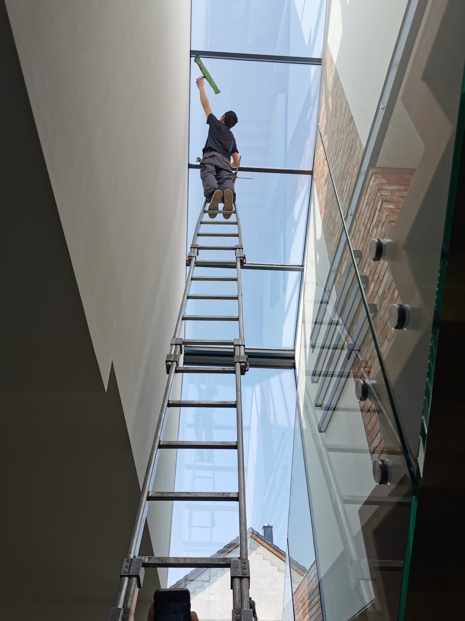 Pracownik na wysokiej drabinie czyści szklany dach wewnątrz budynku, widok z dołu, widoczny budynek na zewnątrz przez szybę.