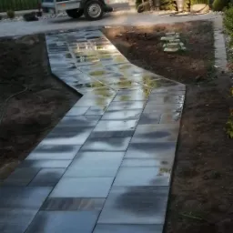 Świeżo ułożony, zakrzywiony chodnik z prostokątnej, szarej kostki brukowej, prowadzący od bramy wjazdowej w głąb posesji, z przyczepką samochodową w tle. Kostka lekko wilgotna, odbija niebo.