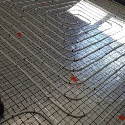 Białe rury ogrzewania podłogowego ułożone na srebrnej folii izolacyjnej z czarną siatką, widoczne pomarańczowe znaczniki i fragment etykiety producenta w rogu.