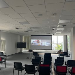 Hilow Solutions - Sala konferencyjna z rzutnikiem sufitowym i ekranem projekcyjnym wyświetlającym prezentację, widoczne krzesła z czerwonymi oparciami oraz wentylacja sufitowa.