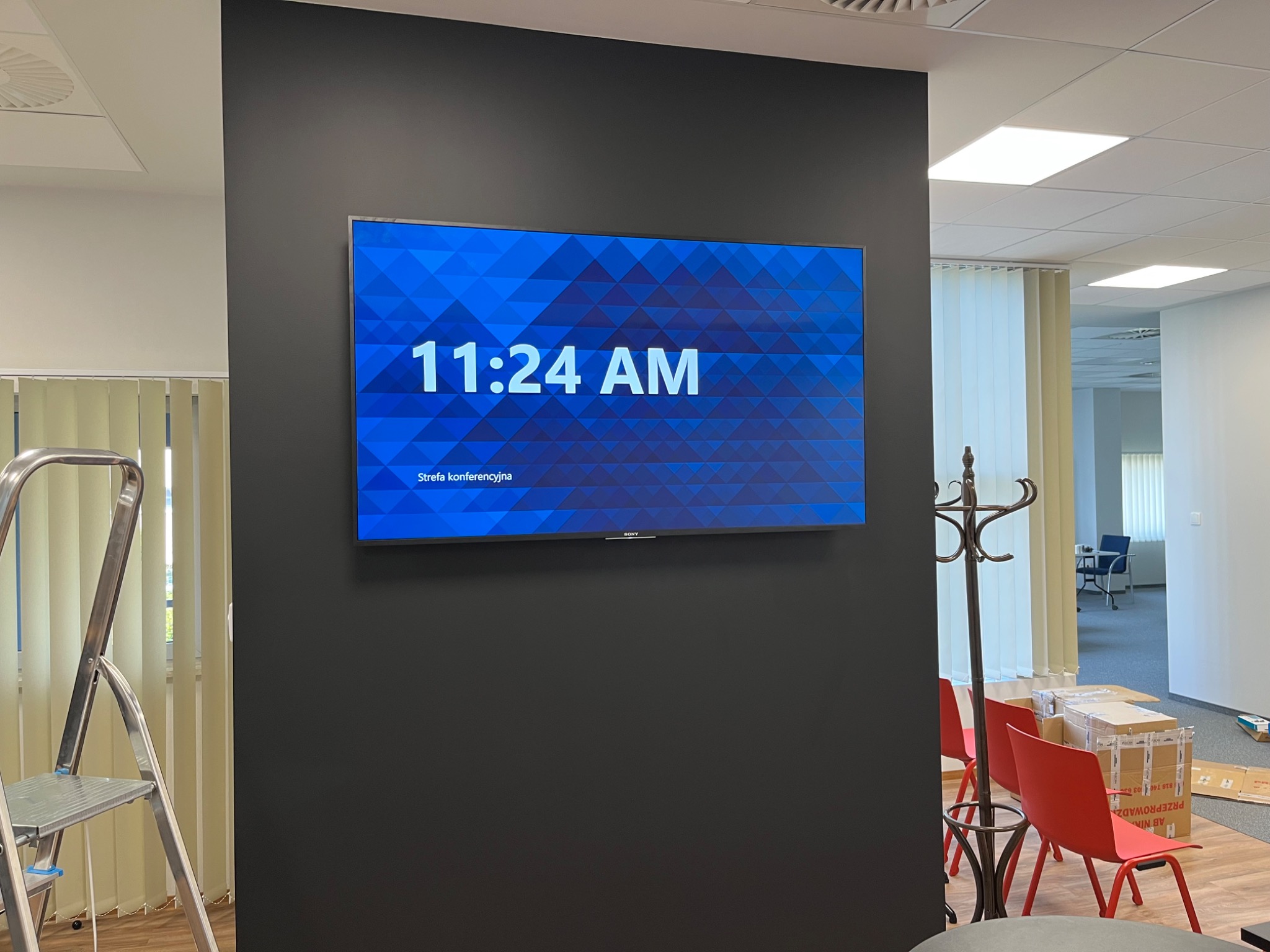Zamontowany telewizor Sony z wyświetlaną godziną 11:24 AM na czarnej ścianie w strefie konferencyjnej, widoczna drabina aluminiowa i krzesła w tle.