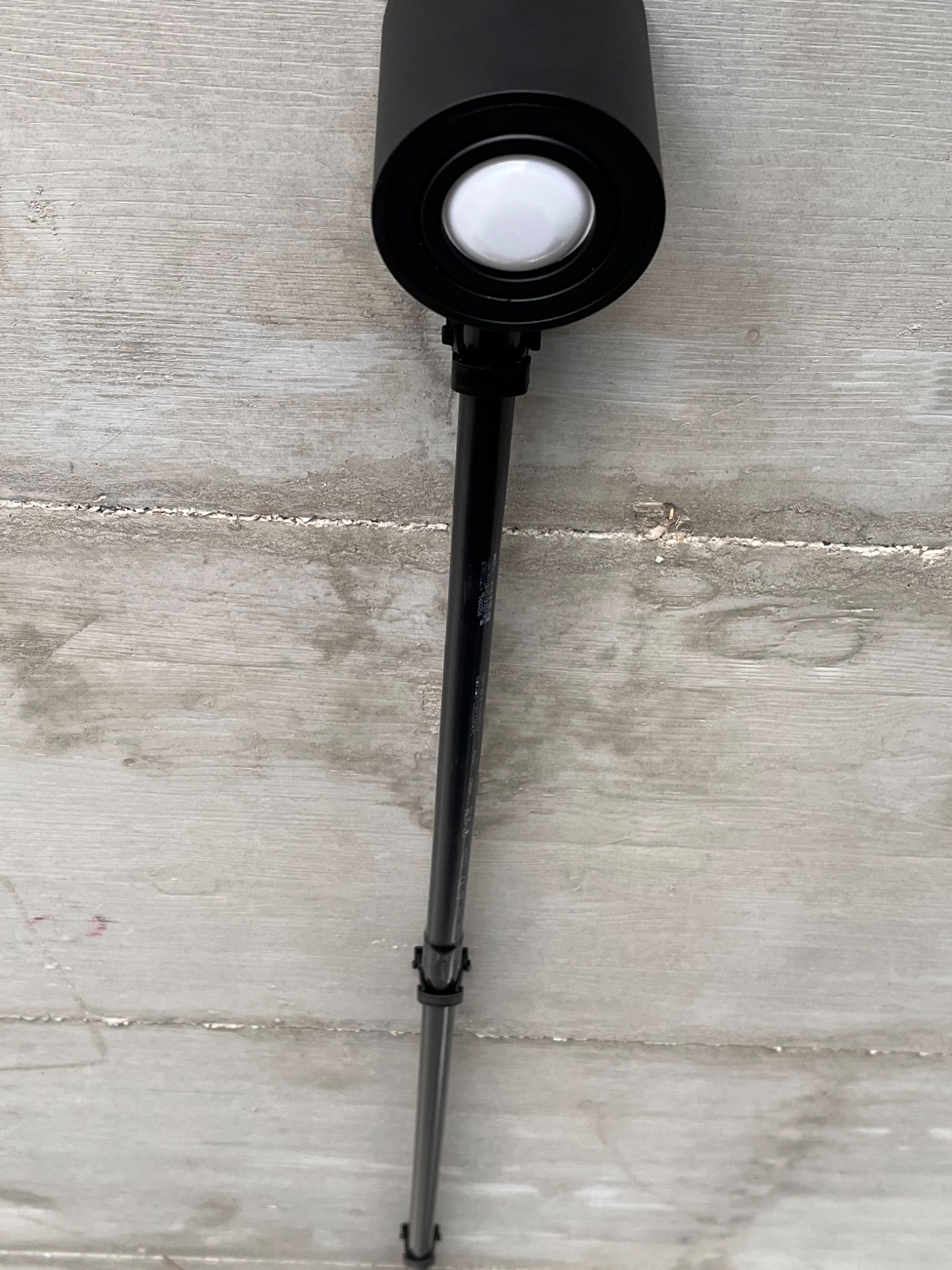 Czarny reflektor LED na regulowanym wysięgniku, zamontowany na szarej, surowej ścianie betonowej z widocznymi śladami szalunku.