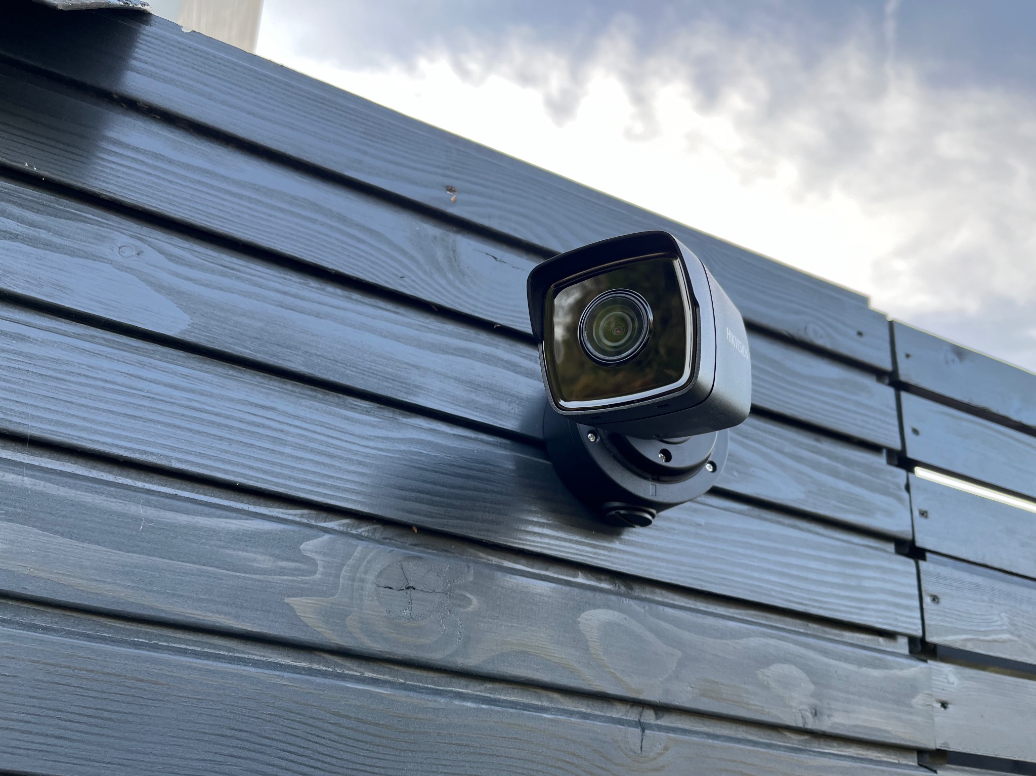 Kamera monitoringu zamontowana na szarej, drewnianej elewacji budynku, widoczna z dołu pod kątem w pochmurny dzień.