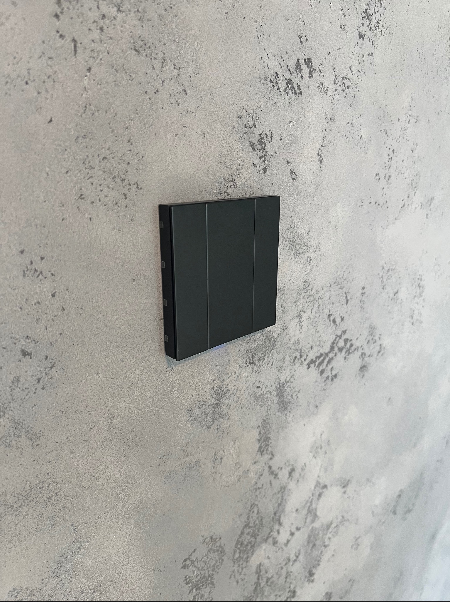 Czarny, nowoczesny panel sterowania o trzech przyciskach zamontowany na szarej ścianie o nieregularnej fakturze.