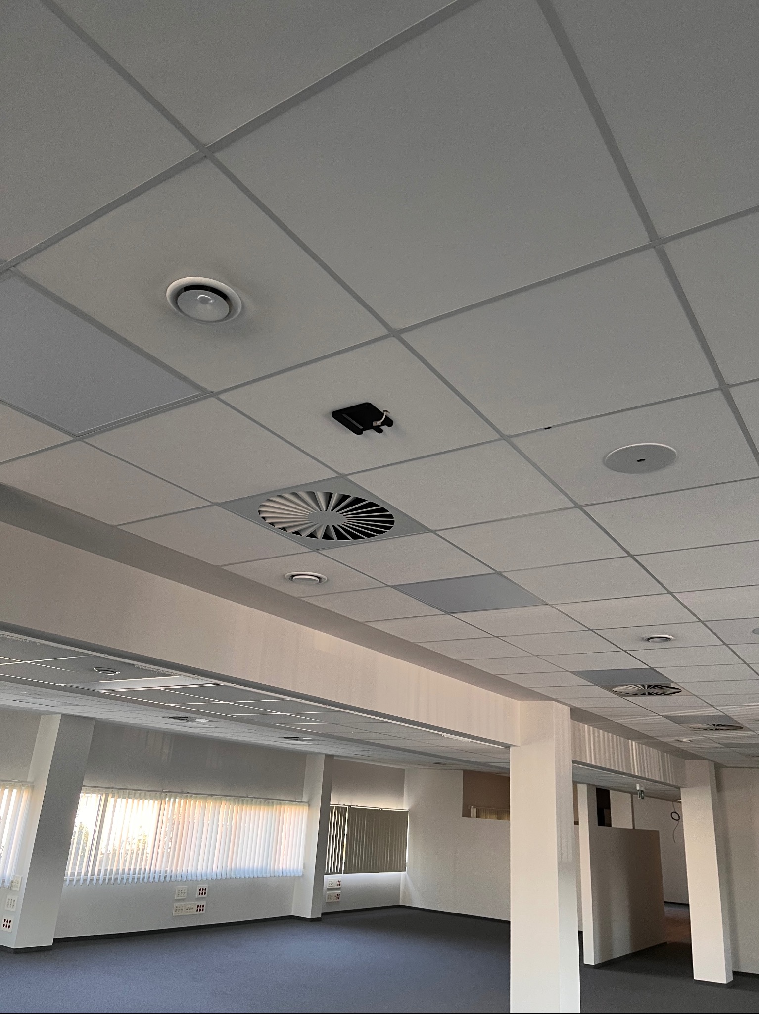Sufit podwieszany w biurze z widocznymi elementami wentylacji, czujnikiem dymu i okrągłymi punktami świetlnymi.