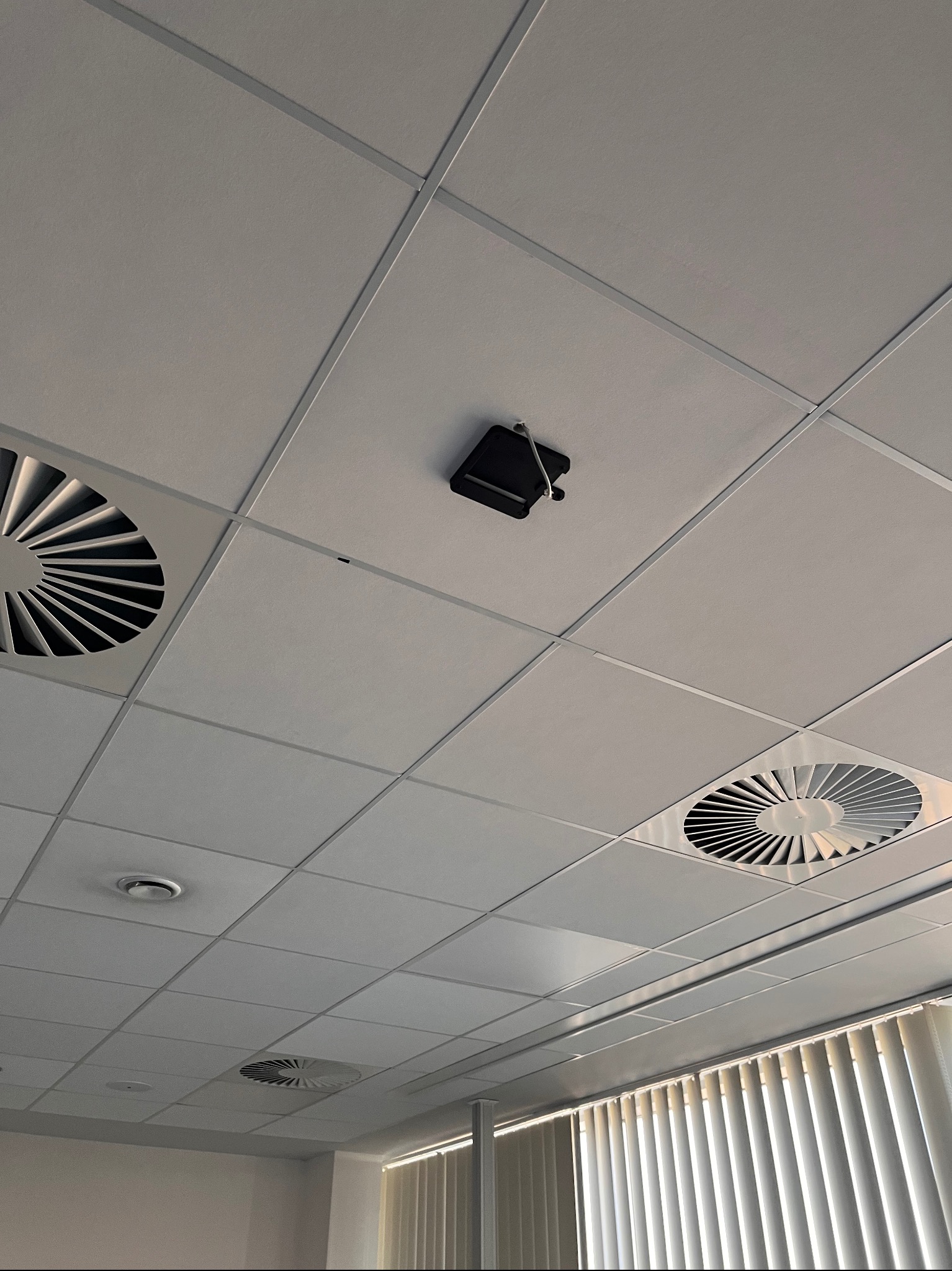 Sufit podwieszany z kratkami wentylacyjnymi i zamontowaną czarną kamerą przemysłową z widocznym kablem.