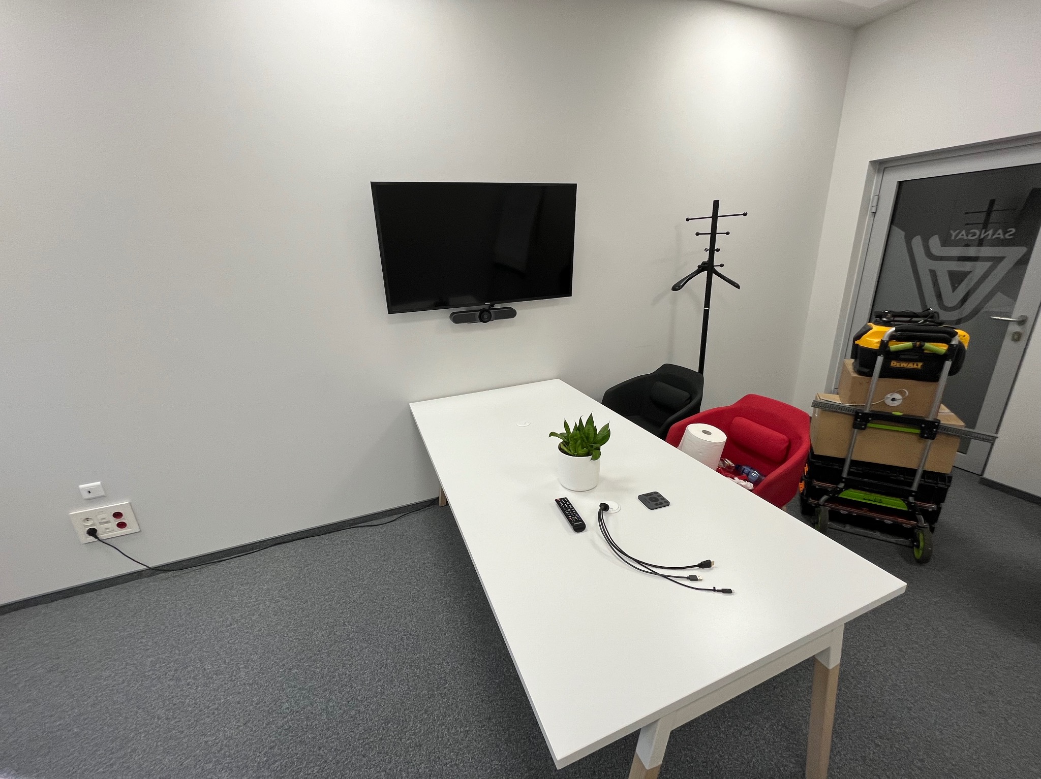 Nowoczesne biuro z zamontowanym telewizorem na ścianie, widoczne gniazdko elektryczne i skrzynka narzędziowa na wózku w tle.