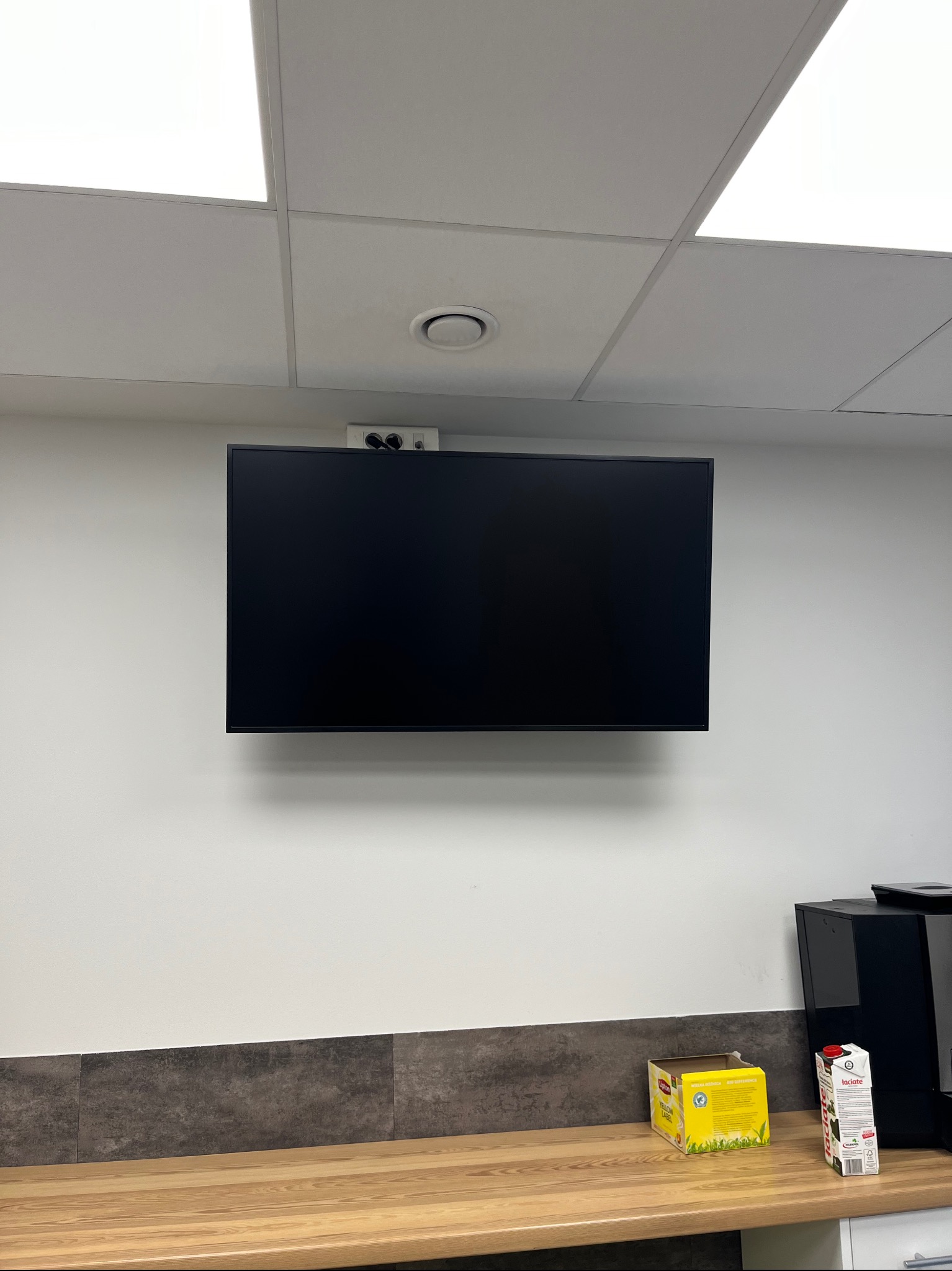 Czarny monitor LCD zamontowany na ścianie biura, nad blatem z pudełkiem herbaty Lipton i kartonem mleka, z widoczną kamerą internetową i głośnikami nad monitorem.