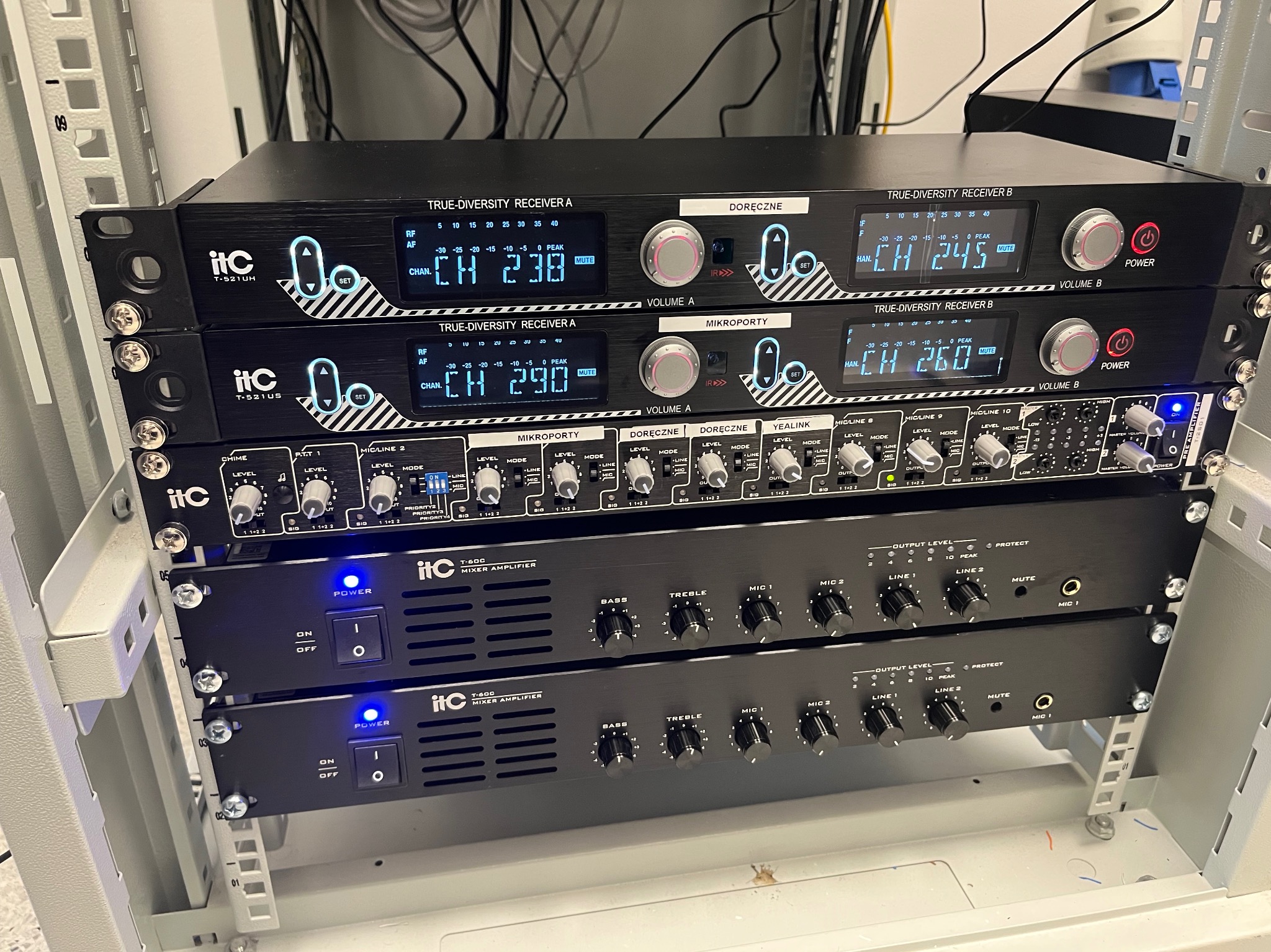 Zestaw czarnych urządzeń audio ITC zamontowanych w metalowej szafie rack, widoczne wyświetlacze, pokrętła i włączniki, kable w tle.