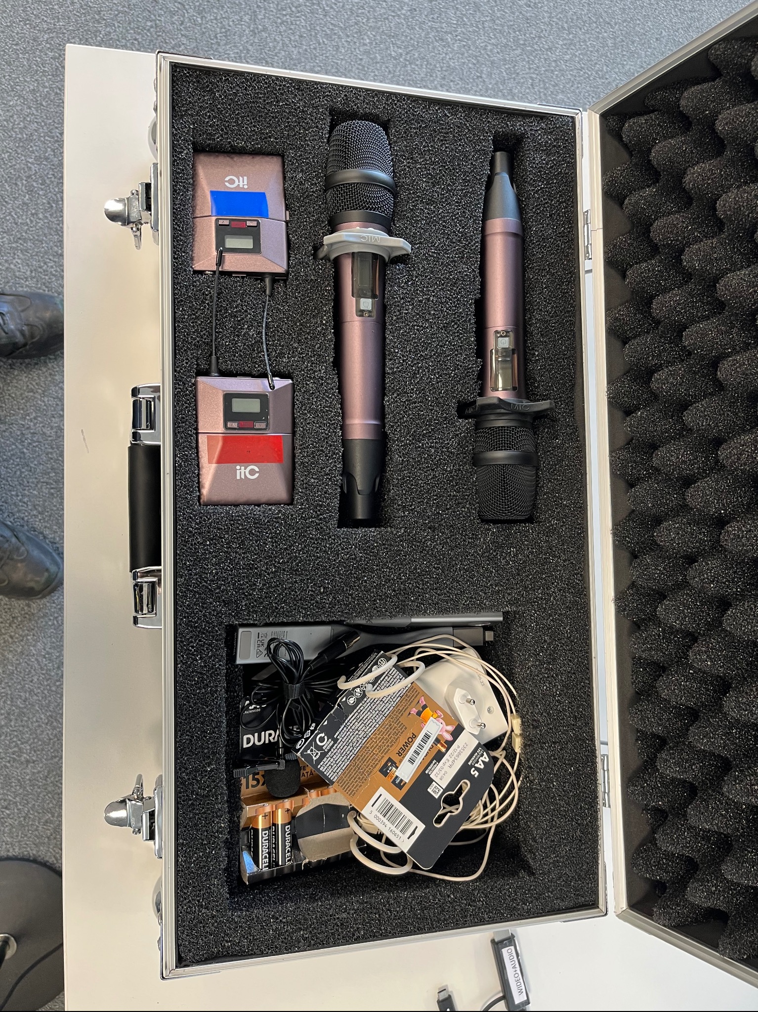 Otwarta aluminiowa walizka z piankowym wypełnieniem zawierająca dwa mikrofony bezprzewodowe w kolorze różowego złota, dwa odbiorniki z wyświetlaczami, baterie Duracell, zasilacz i kable.