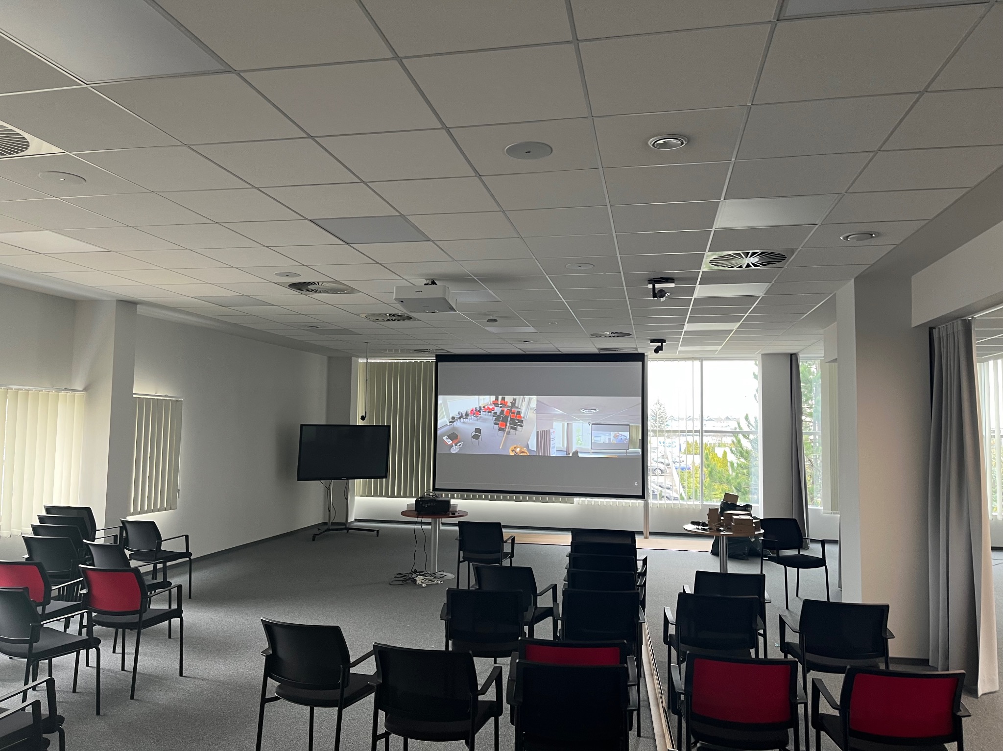 Sala konferencyjna z rzutnikiem sufitowym i ekranem projekcyjnym wyświetlającym prezentację, widoczne krzesła z czerwonymi oparciami oraz wentylacja sufitowa.